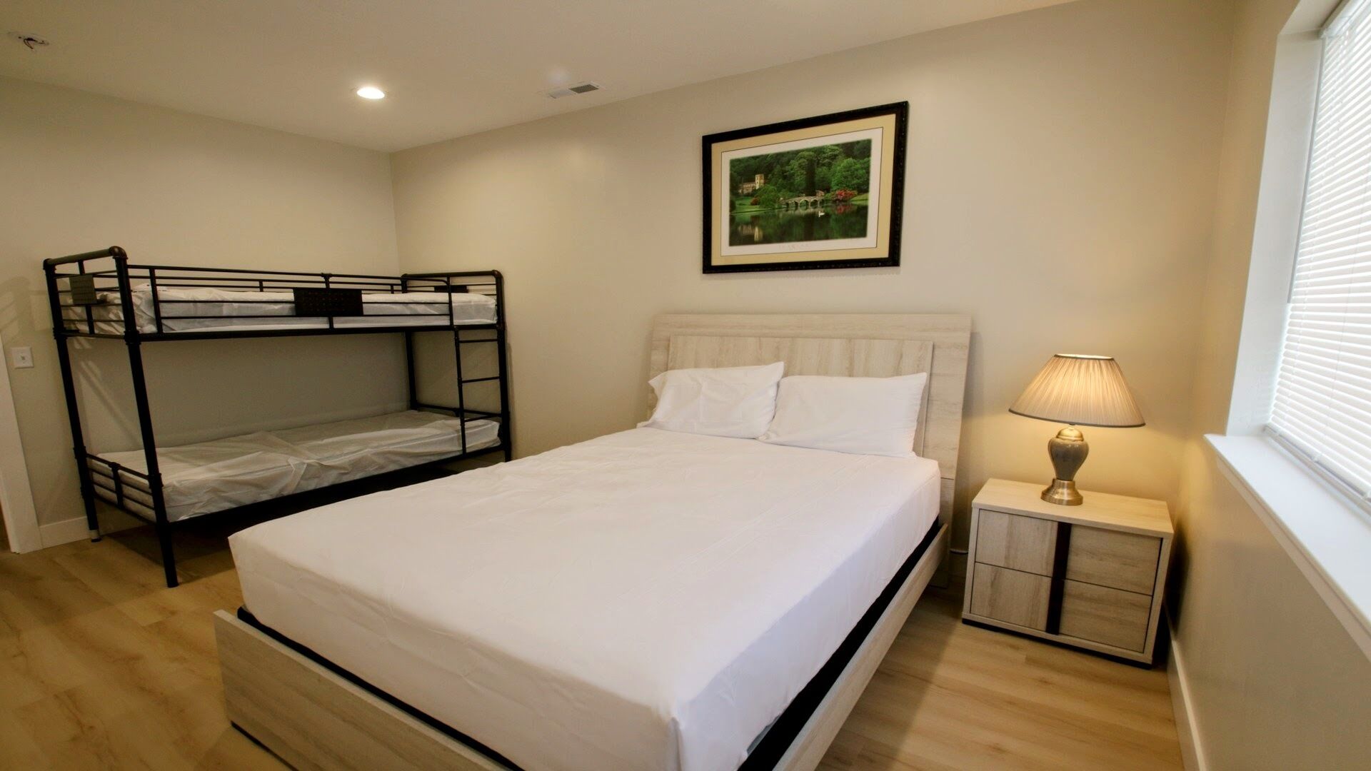 A bedroom with a bunk bed, a single bed, a bedside table with a lamp, and a framed picture on the wall.