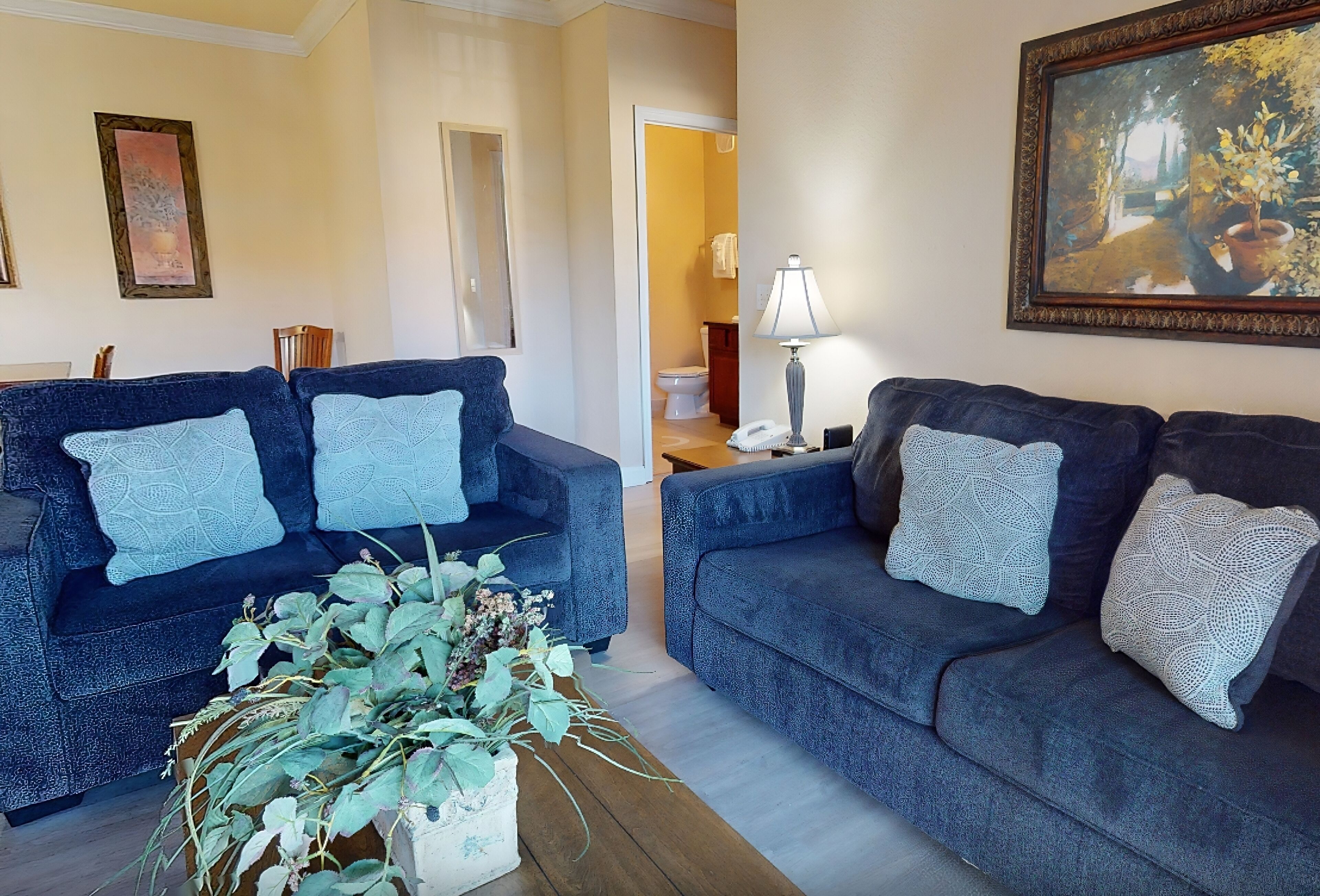 A living room with two dark blue sofas, each with a white knitted pillow. There is a wooden coffee table with a potted plant in the center. A painting is on the wall above the right sofa.