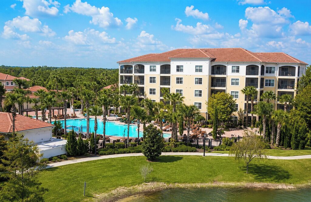 A multi-story hotel with a swimming pool, surrounded by palm trees and a grassy area.