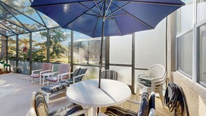 Uma sala de vidro com uma mesa redonda e cadeiras, um grande guarda-sol azul, e vista para árvores e um edifício ao fundo.