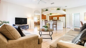 A spacious living room with a sofa, a television, and a dining area with wooden cabinets and a kitchen in the background. - Gorgeous home near Disney with private pool, pool spa, on-site gym, tennis, AC (Davenport)