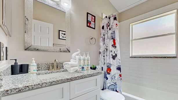 Une salle de bain avec des toilettes blanches, un lavabo doté d’un plan de travail à motifs, un miroir avec un cadre décoratif et un rideau de douche orné de personnages de dessins animés.