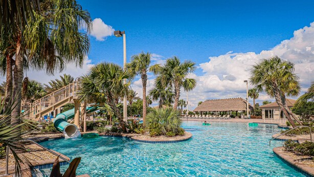 Ein Poolbereich eines Resorts mit Wasserrutsche, Palmen und einem Gebäude mit Reetdach.