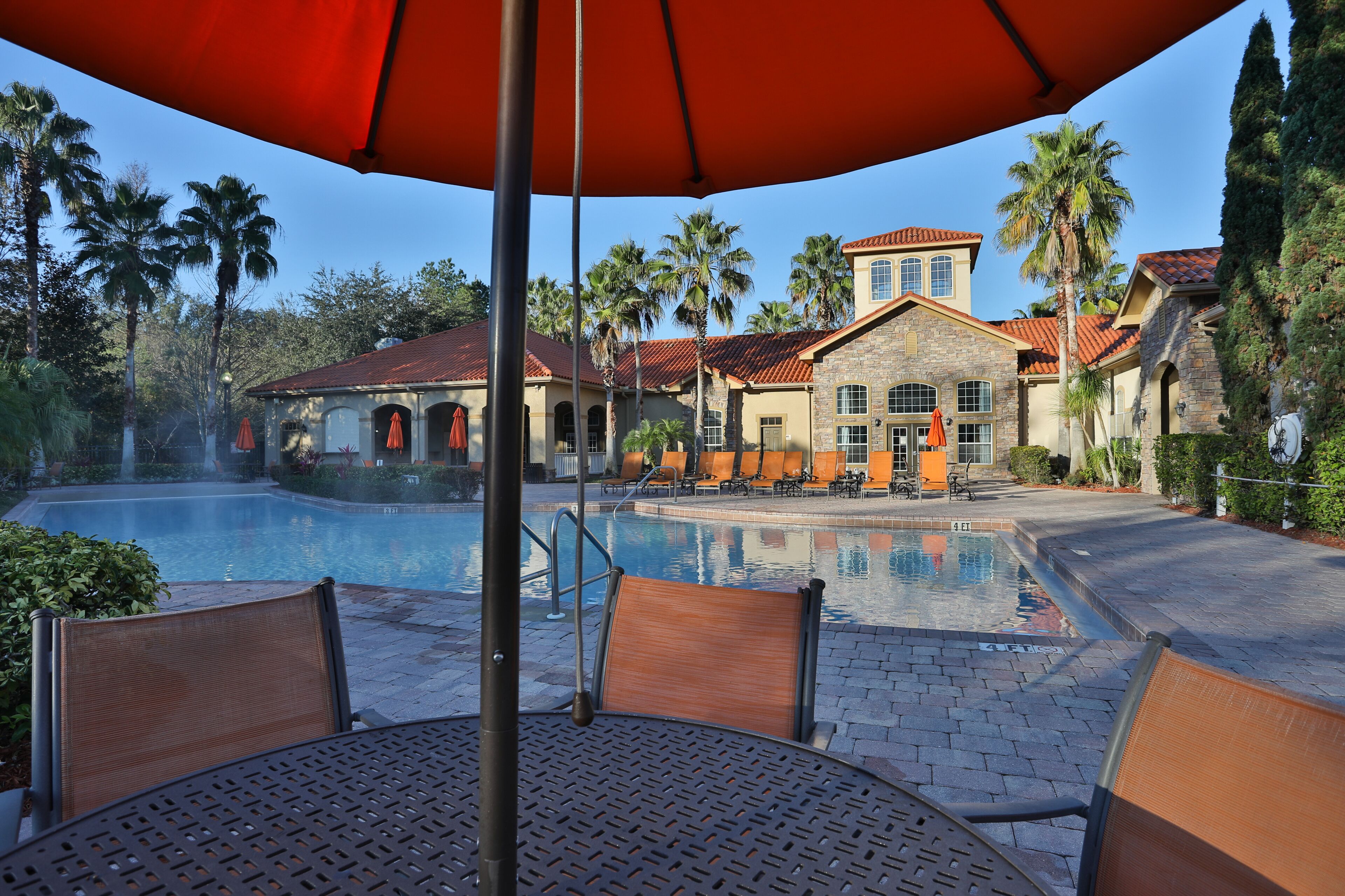 A pool area with lounge chairs and umbrellas, surrounded by palm trees and a building with a tiled roof.