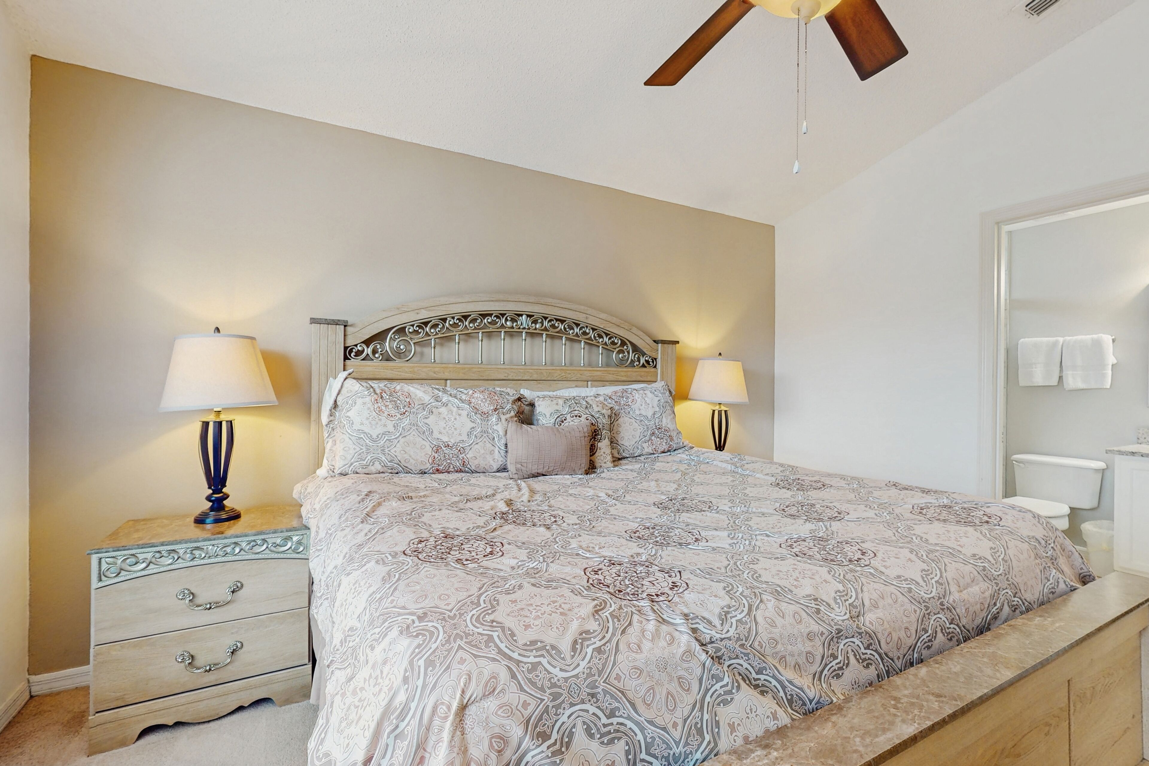 A bedroom with a bed, two bedside tables with lamps, a ceiling fan, and a bathroom visible through an open door.