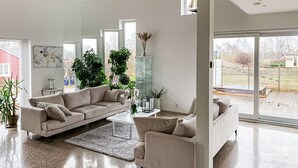 Moderni olohuone, jossa on beigenvärinen sohva, sohvapöytä ja lasikaappi. Huoneessa on suuret ikkunat, joista on näkymä ulos.