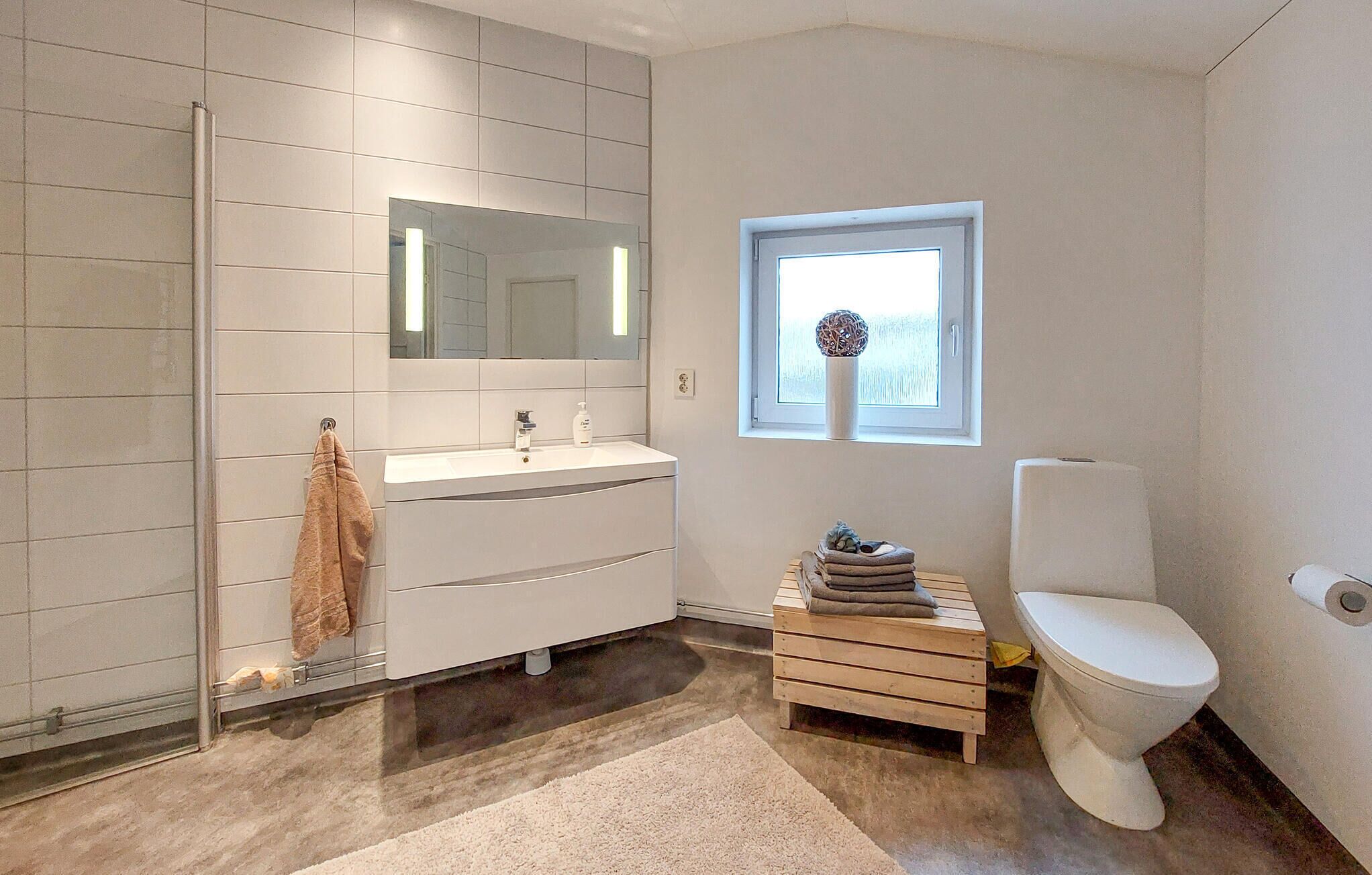 A modern bathroom with a glass shower enclosure, a white sink with a mirror above, a toilet, and a small wooden stool with towels.
