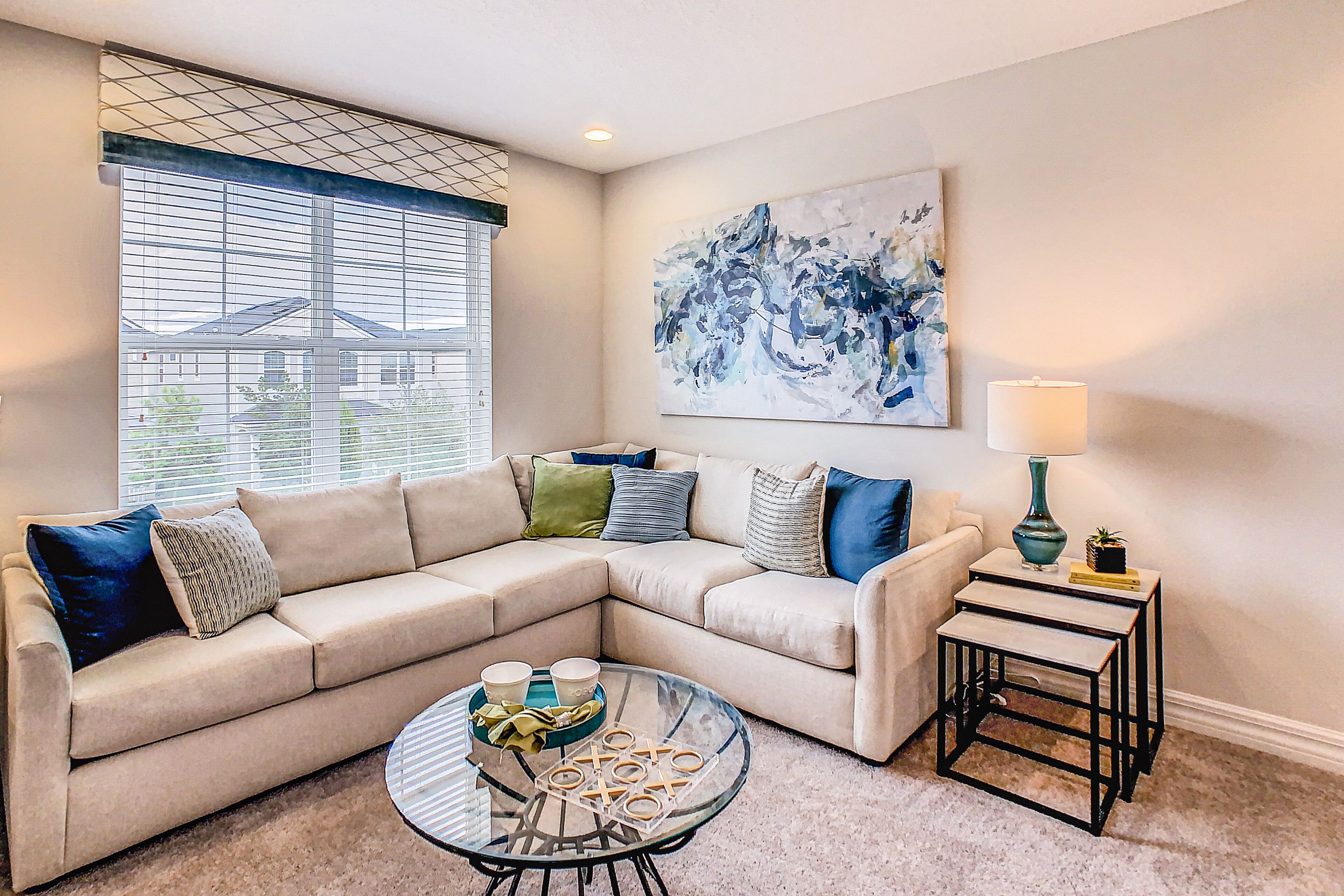 A living room with a beige sectional sofa, a glass coffee table with board games, a blue abstract painting, and a lamp on a side table.