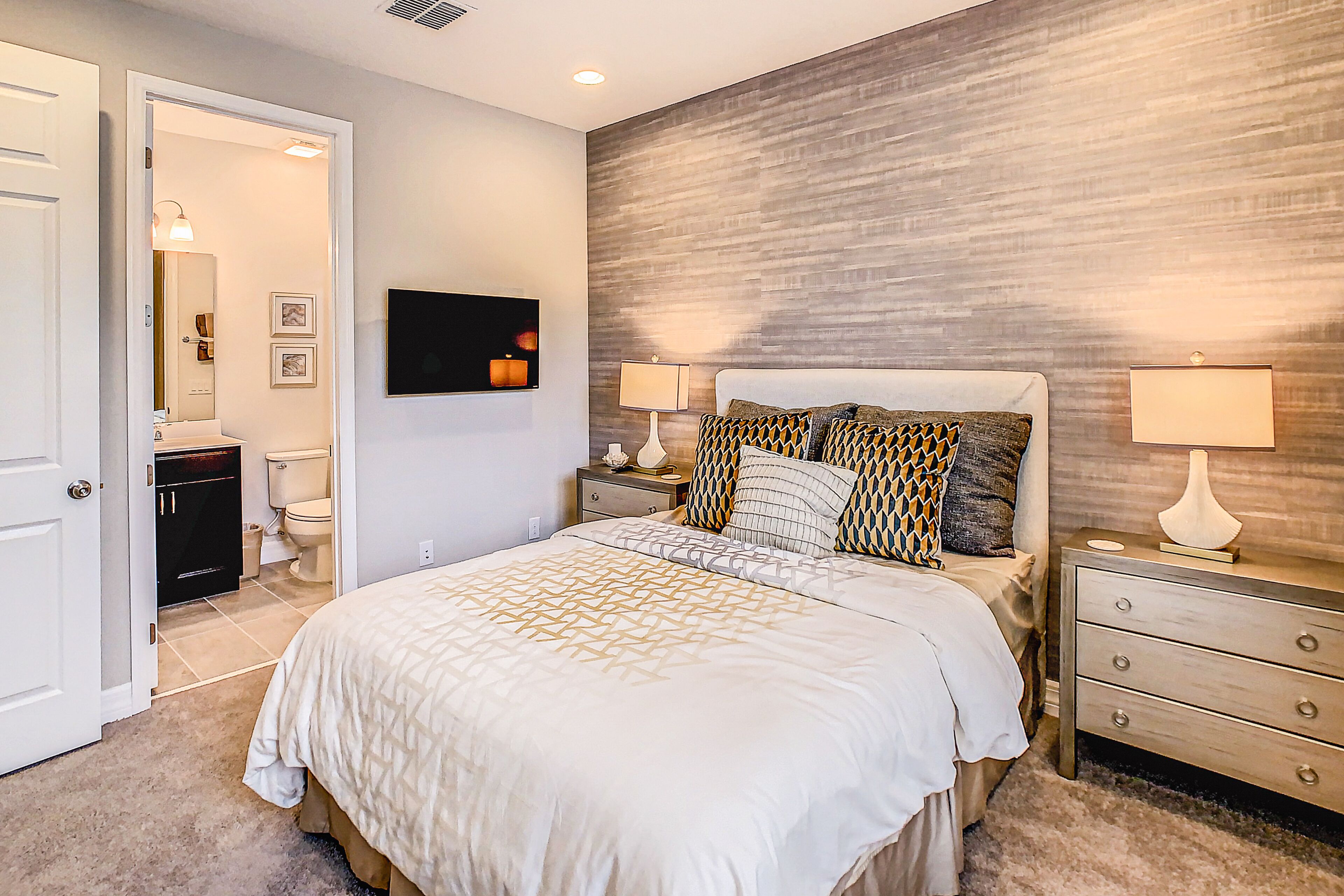 Une chambre à coucher avec un grand lit, deux tables de chevet avec des lampes, une tête de lit en bois et une salle de bain visible à travers une porte ouverte.