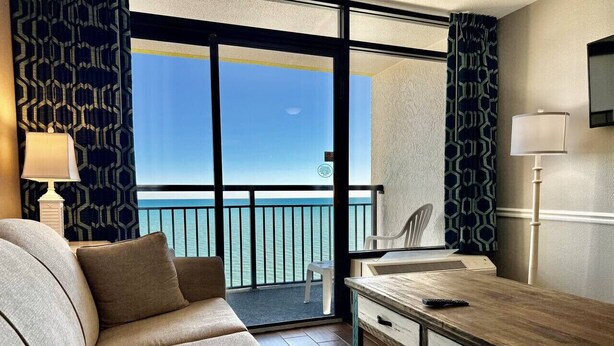 Une pièce comprenant un canapé, un bureau en bois, une chaise et offrant une vue sur la mer grâce à de grandes fenêtres.