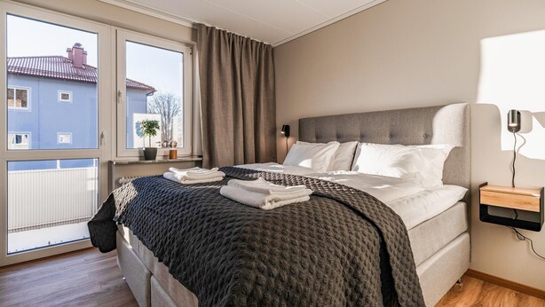 A bedroom with a bed, a bedside table, a lamp, a window with curtains, and a view of buildings outside. - 1 bedroom nice apartment in Huskvarna (Huskvarna)