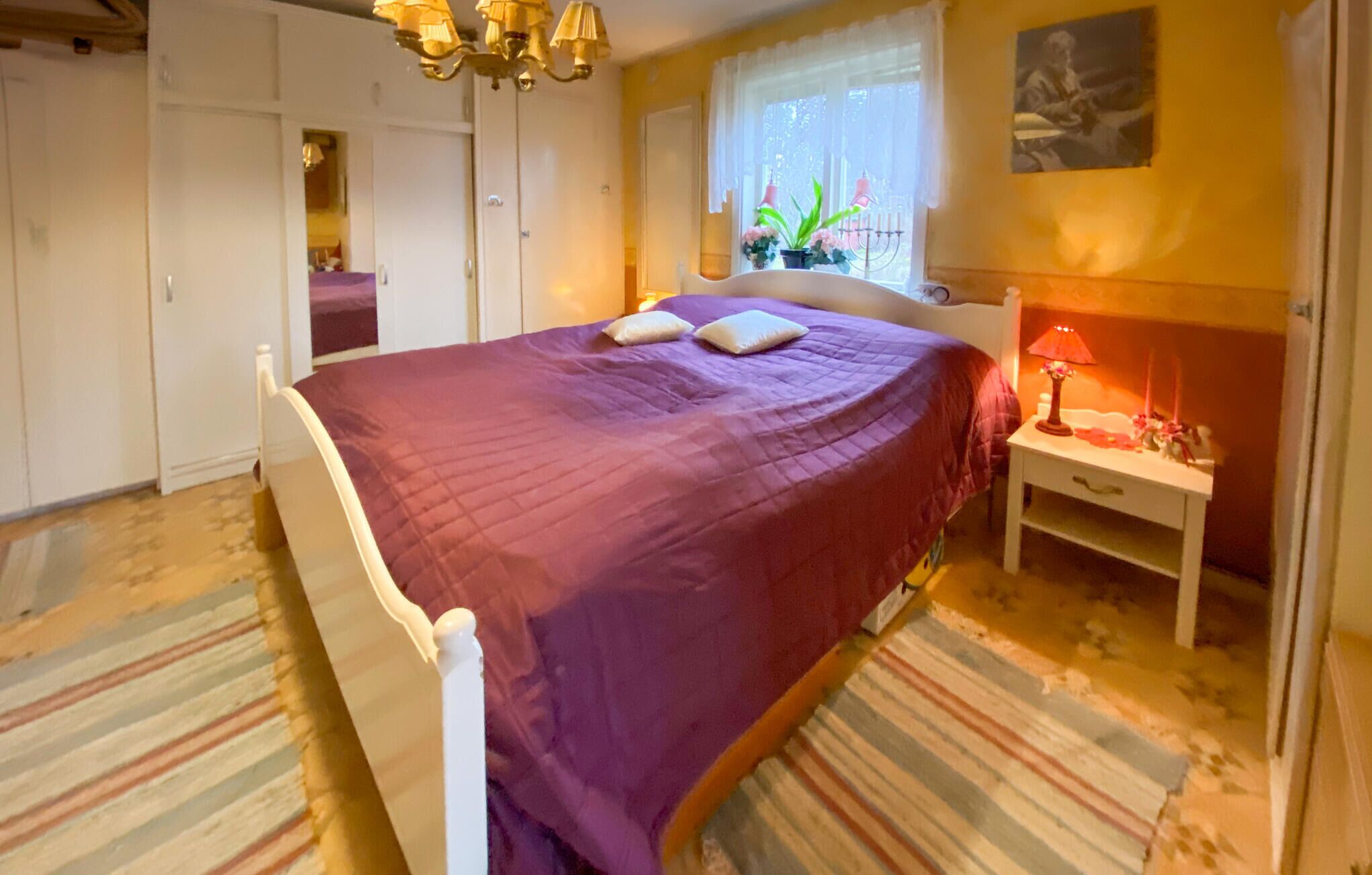 A bedroom with a bed, a nightstand, a window with plants, and a painting on the wall.