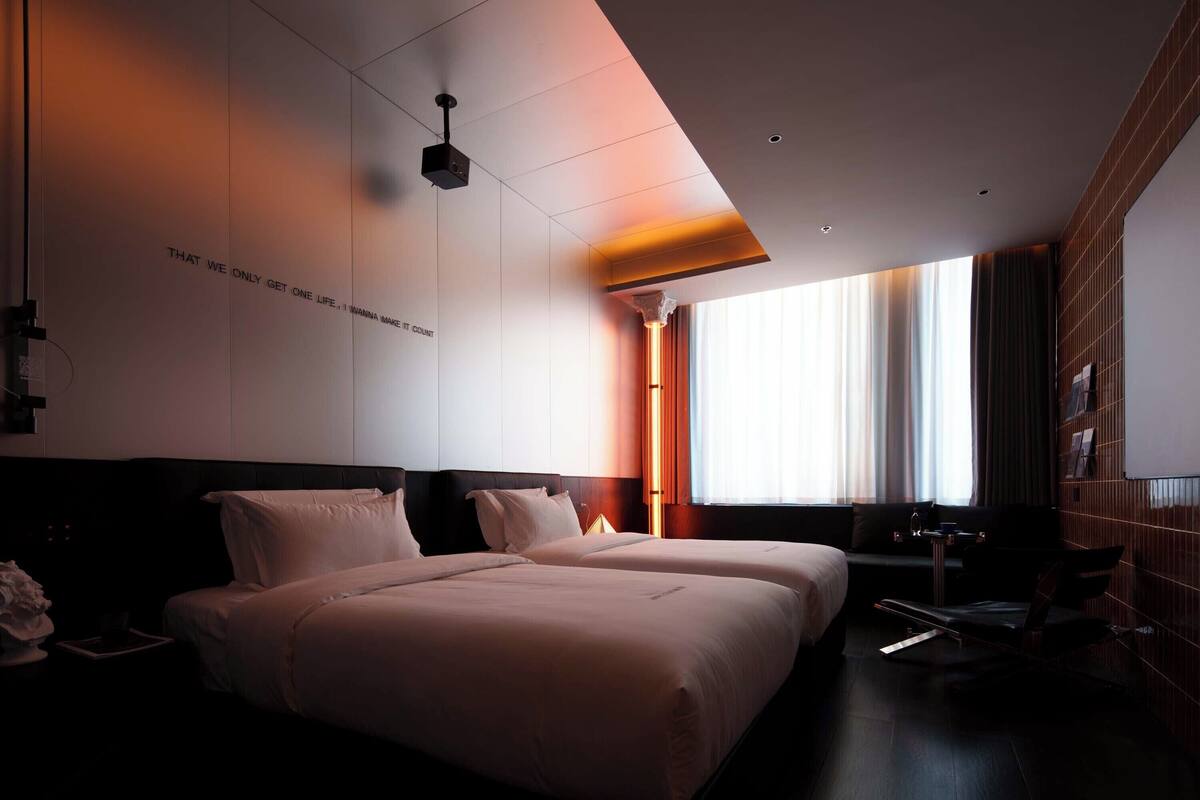 Et moderne hotelværelse med to senge, et skrivebord og et stort vindue med gardiner.