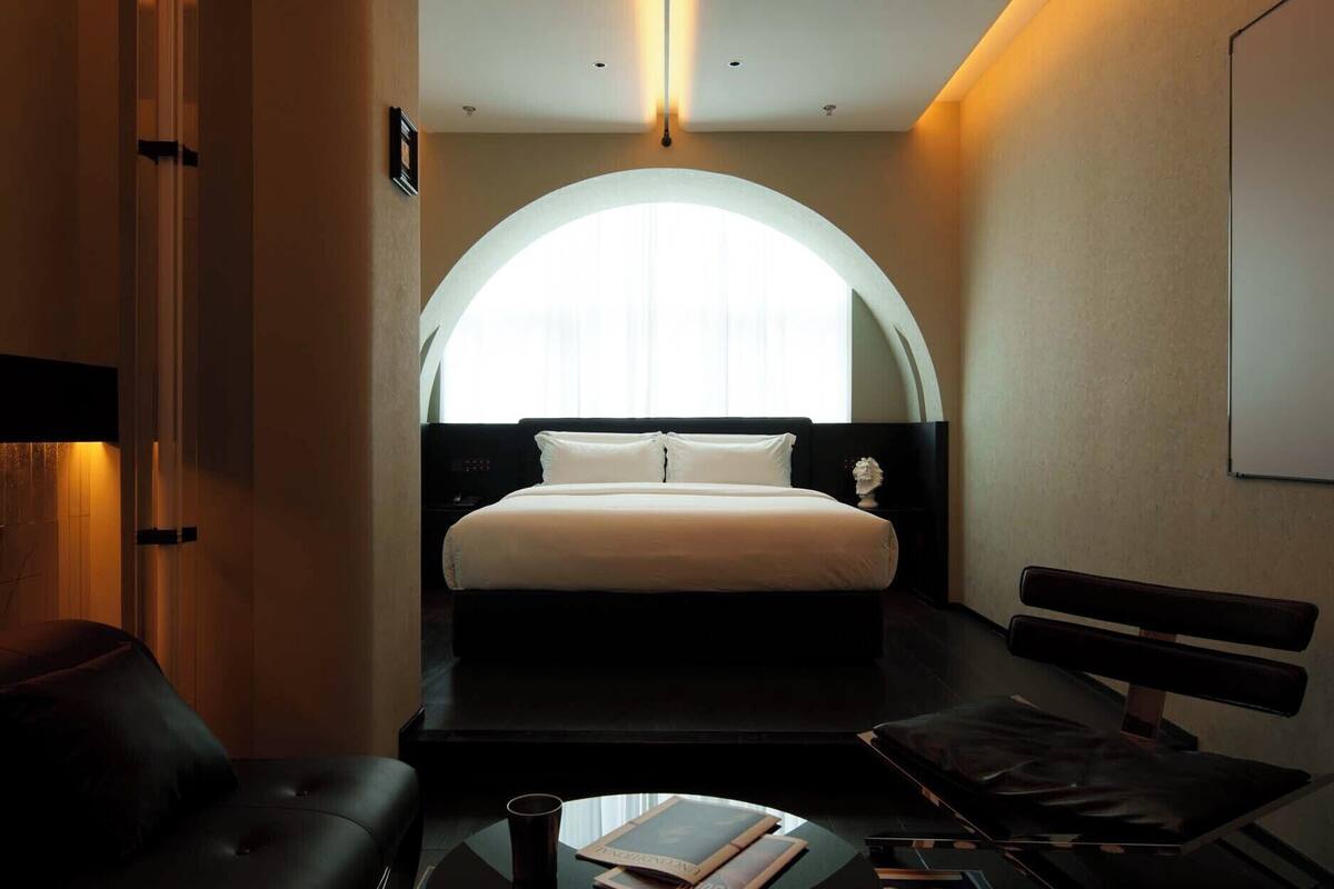 Et moderne hotelværelse med en stor seng, et rundt bord, en sofa og udsigt over byen gennem et stort, hvælvet vindue.