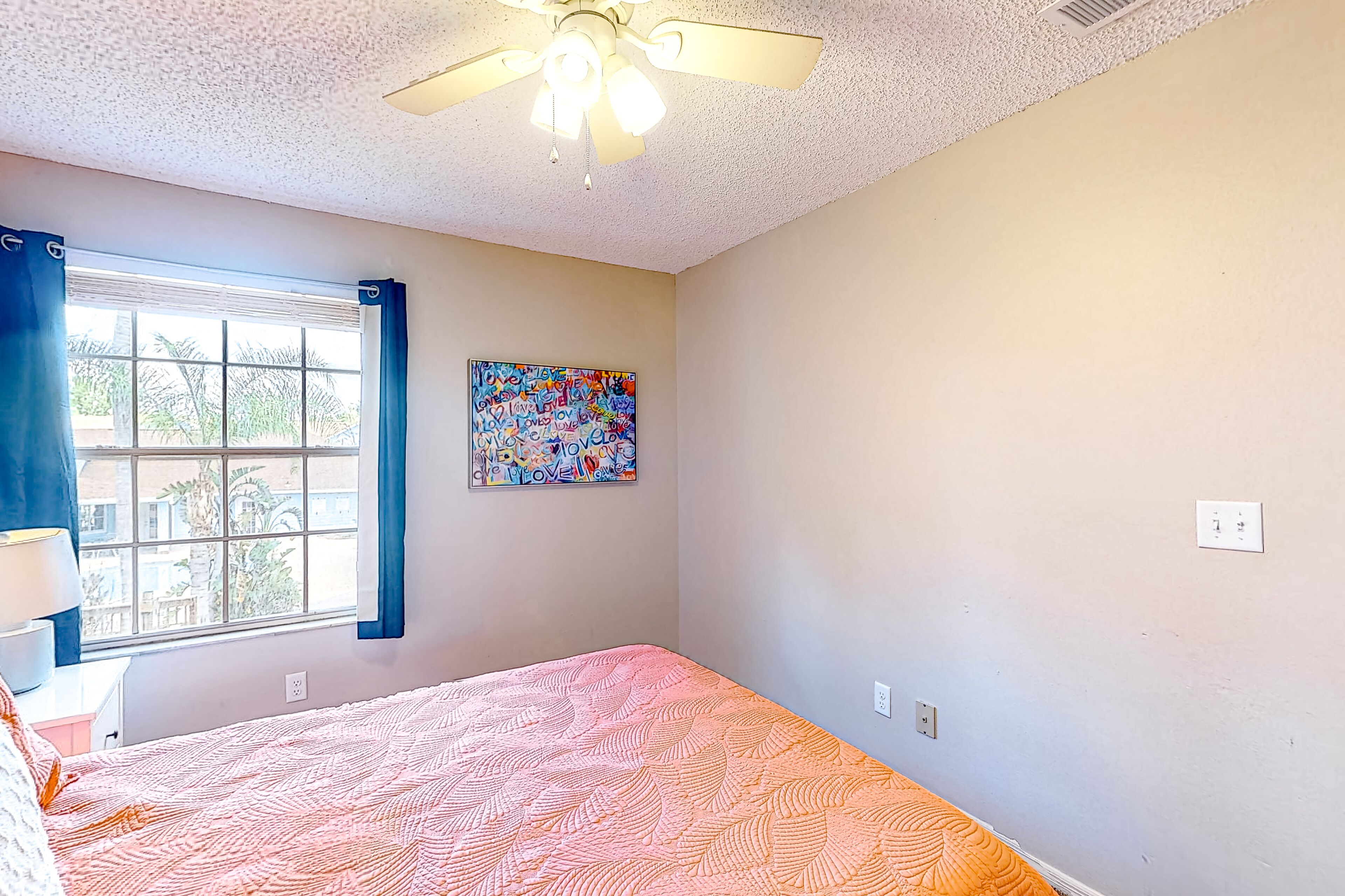 A bedroom with a bed, a ceiling fan, a window with curtains, and a colorful artwork on the wall.