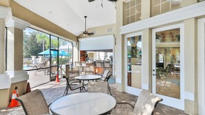 Un espace extérieur aménagé en terrasse, avec des tables et des chaises rondes, un ventilateur de plafond et de grandes fenêtres donnant sur une piscine.