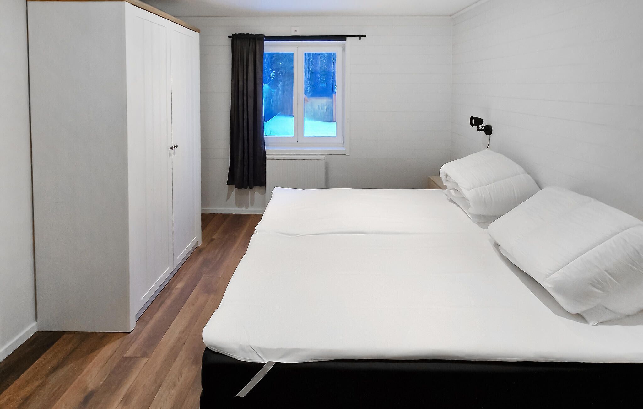 A bedroom with a bed, a window with curtains, a wardrobe, and a bedside table with a lamp.