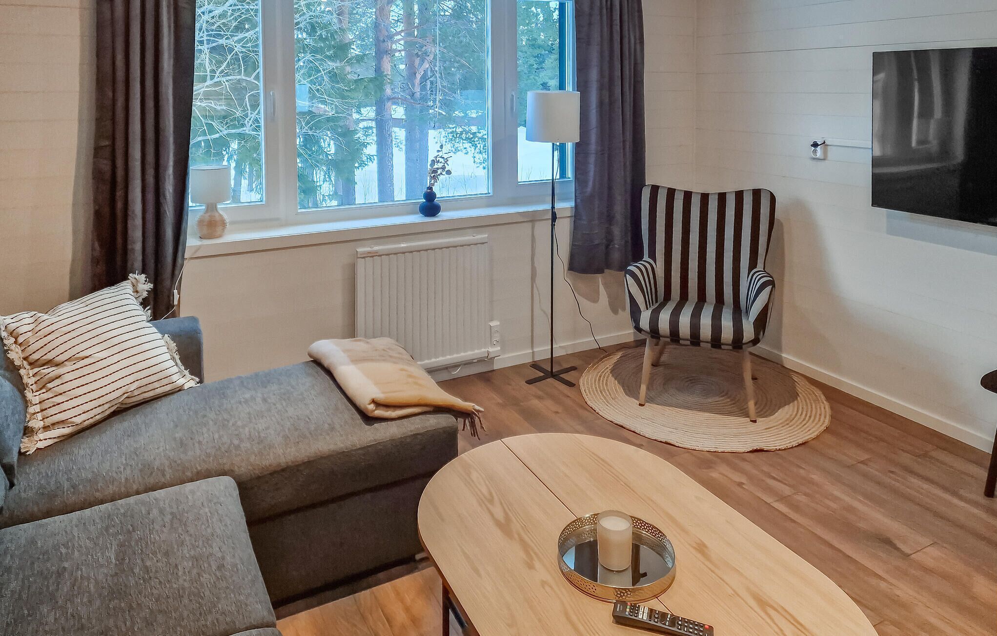 A cozy living room with a sofa, a striped armchair, a round coffee table with a candle, and a view of trees through the window.
