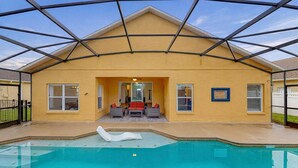 Ein überdachter Außenpool mit einem klaren, blauen Wasserspiel, eine Terrasse mit Sitzgelegenheiten und ein gelbes Gebäude mit Giebeldach.