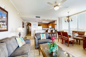 Ein geräumiges Wohnzimmer mit einer grauen Couch, einem Glastisch und einem Essbereich mit Holzsesseln und einem Glastisch. Im Wohnzimmer sind ein Deckenventilator und eine Deckenleuchte zu sehen.