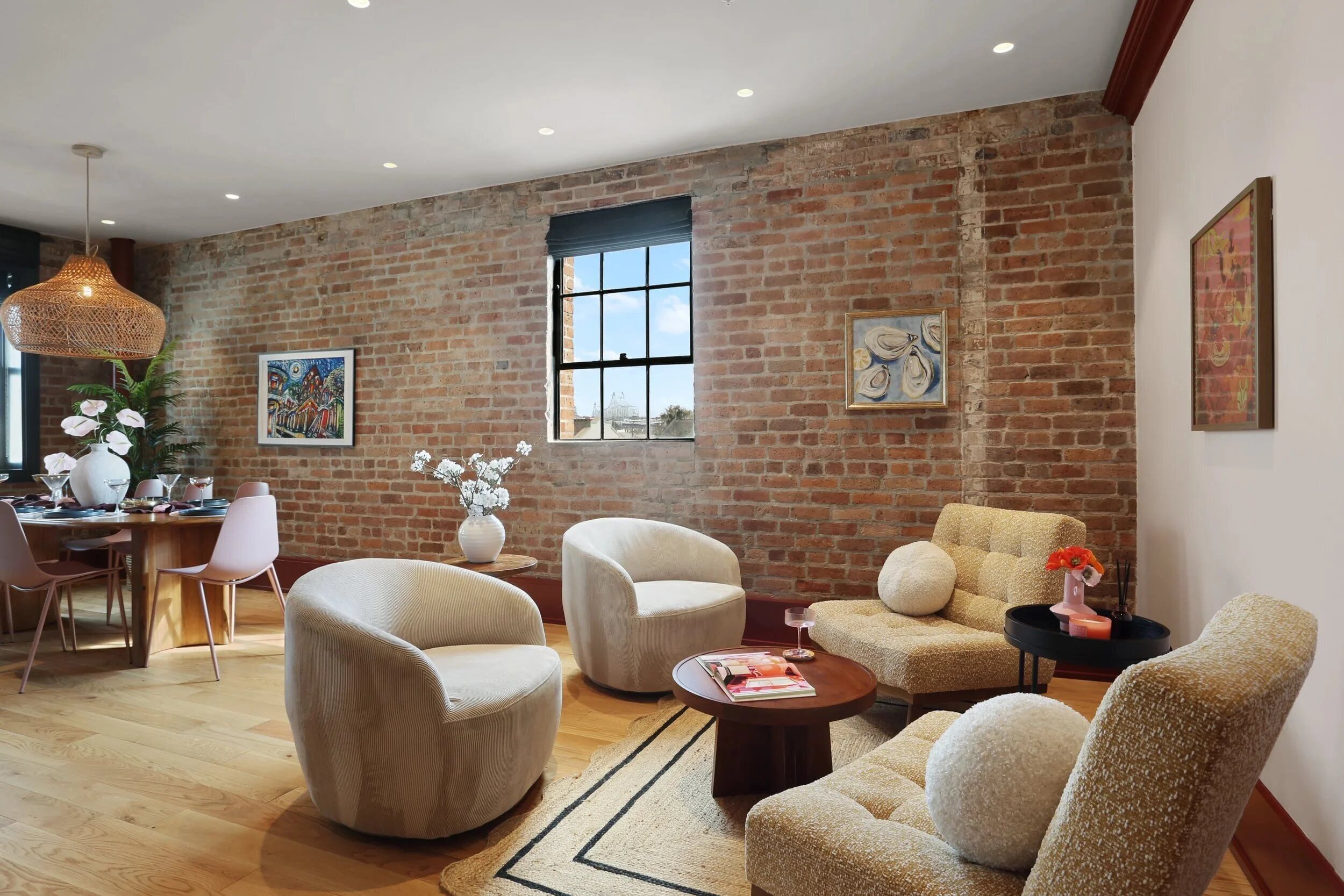 A spacious living room with a brick wall, a dining area with a wooden table and chairs, and a large window providing natural light.