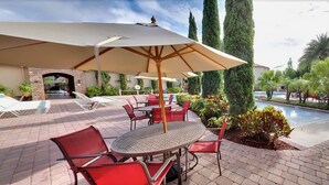 Une terrasse extérieure avec des chaises rouges et une table placée sous un grand parasol, à proximité d’une piscine entourée de verdure.