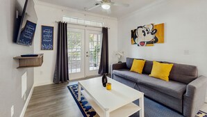 Un salon avec un canapé gris, des coussins jaunes, une table basse blanche, un ventilateur de plafond et une grande fenêtre avec des rideaux.