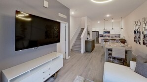 Un salon moderne doté d’un téléviseur à écran plat, d’un escalier menant à un espace repas et d’une cuisine en arrière-plan.