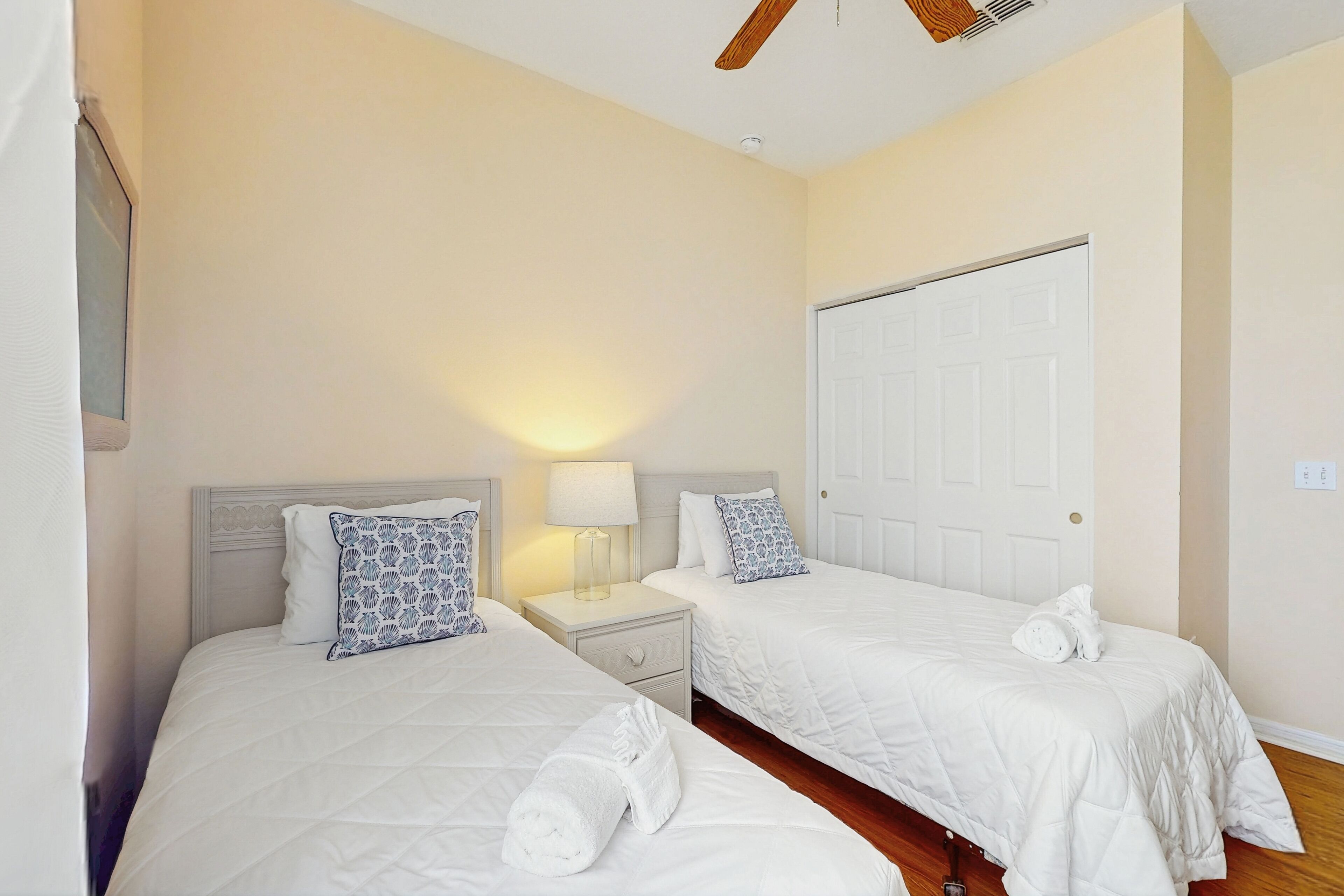 A hotel room with two beds, a nightstand with a lamp, a wooden ceiling fan, and a white door.