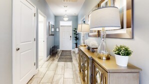 Un pasillo con pared azul claro, un mueble de madera con una lámpara, una planta en maceta y una puerta al final.