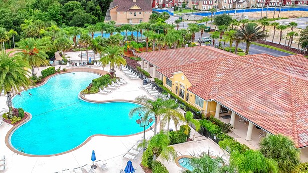 Uma piscina grande cercada por espreguiçadeiras e palmeiras, com um edifício de telhado vermelho ao fundo.