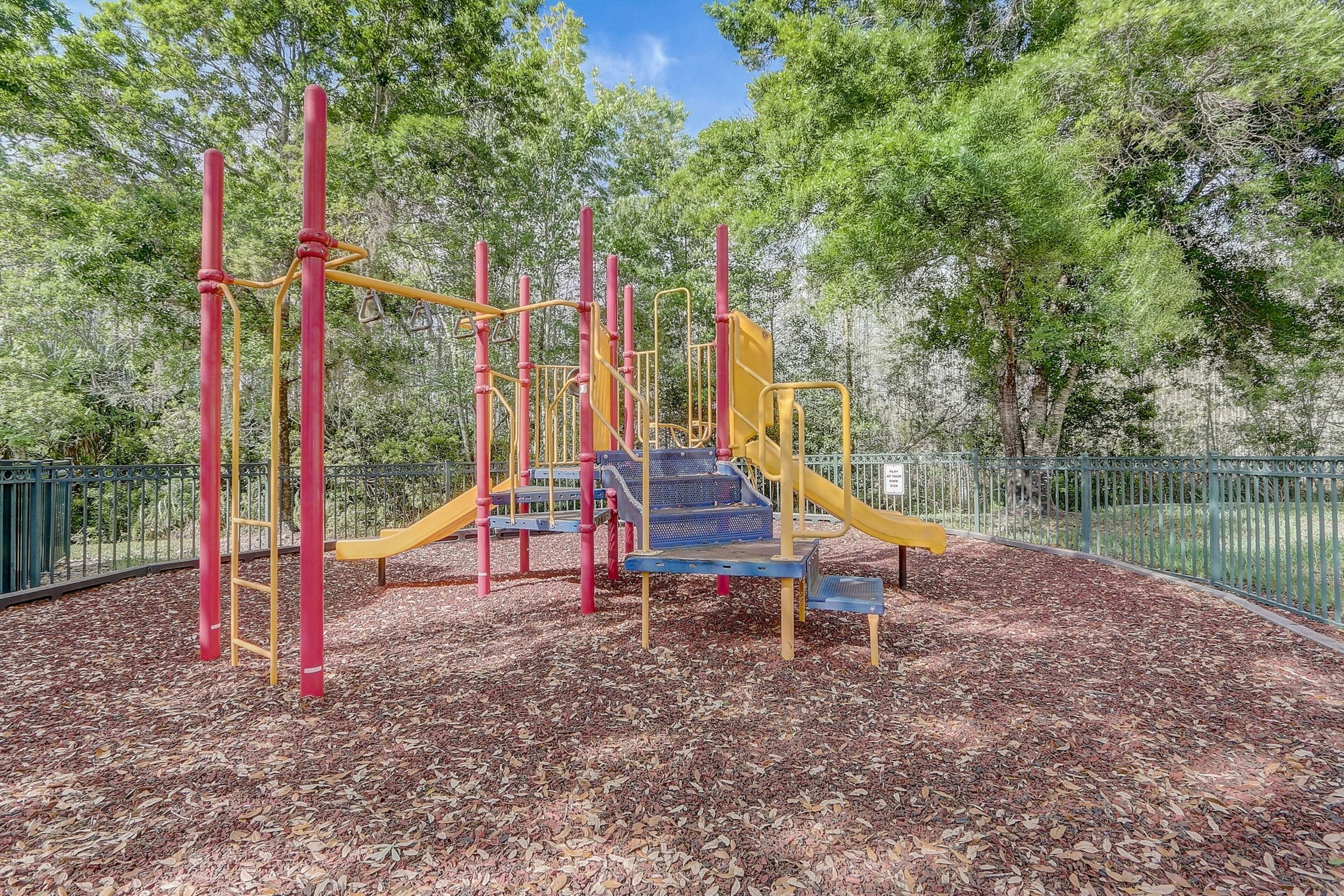 A playground with climbing structures and slides, surrounded by a fence and trees.