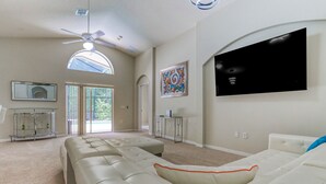 Un salón espacioso con un sofá seccional grande, un ventilador de techo, un televisor de pantalla plana montado en la pared y un ventanal que proporciona luz natural.