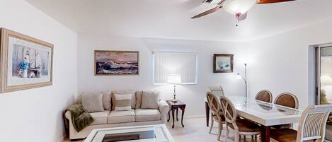 A living room with a sofa, coffee table, dining table, chairs, ceiling fan, and artwork on the walls.