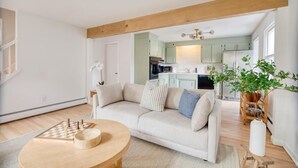 Een moderne woonkamer met een beige bank, een houten salontafel en een schaakbord. In de achtergrond is een keuken zichtbaar met lichtgroene kastjes en een witte koelkast.