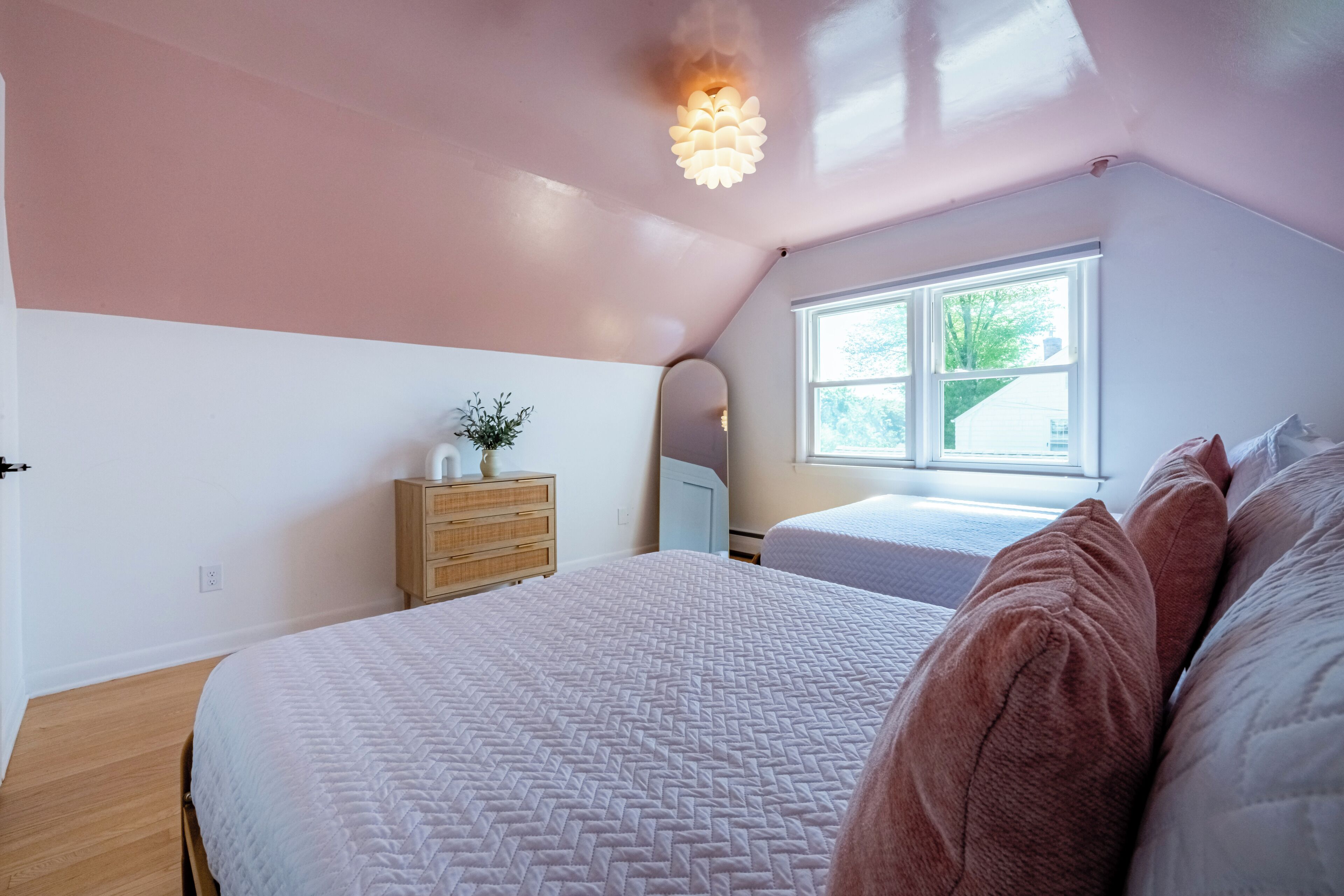 A bedroom with a large bed, a dresser, a window with a view of greenery, and a ceiling light fixture.