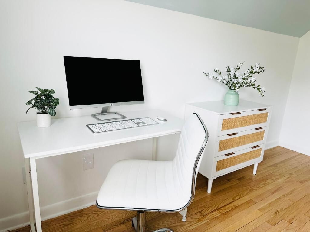 Un bureau blanc avec un ordinateur, une chaise blanche, une petite plante et une commode blanche avec des tiroirs.