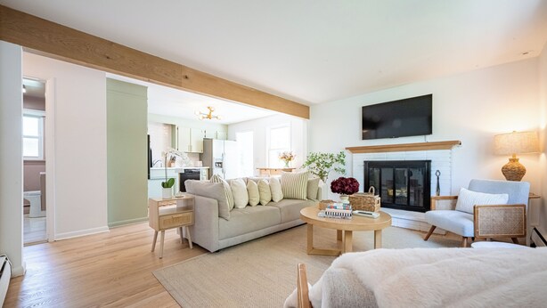 Een moderne woonkamer met een bank, salontafel en stoelen. Er is een open haard, een aan de muur bevestigde televisie en een keuken zichtbaar op de achtergrond.