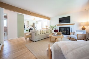 A modern living room with a sofa, coffee table, and chairs. There is a fireplace, a television mounted on the wall, and a kitchen visible in the background. - Cozy Cottage with Fire Pit (Fairfield)