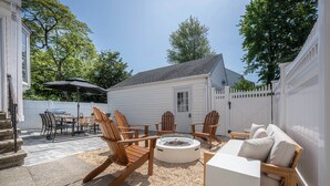 Een achtertuin met een vuurkuil, houten stoelen, een witte bank en een eettafel met stoelen.
