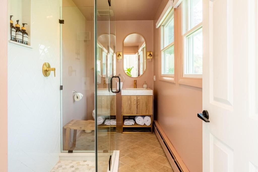 A bathroom with a glass shower enclosure, a wooden bench, a sink with a mirror, and a towel rack.