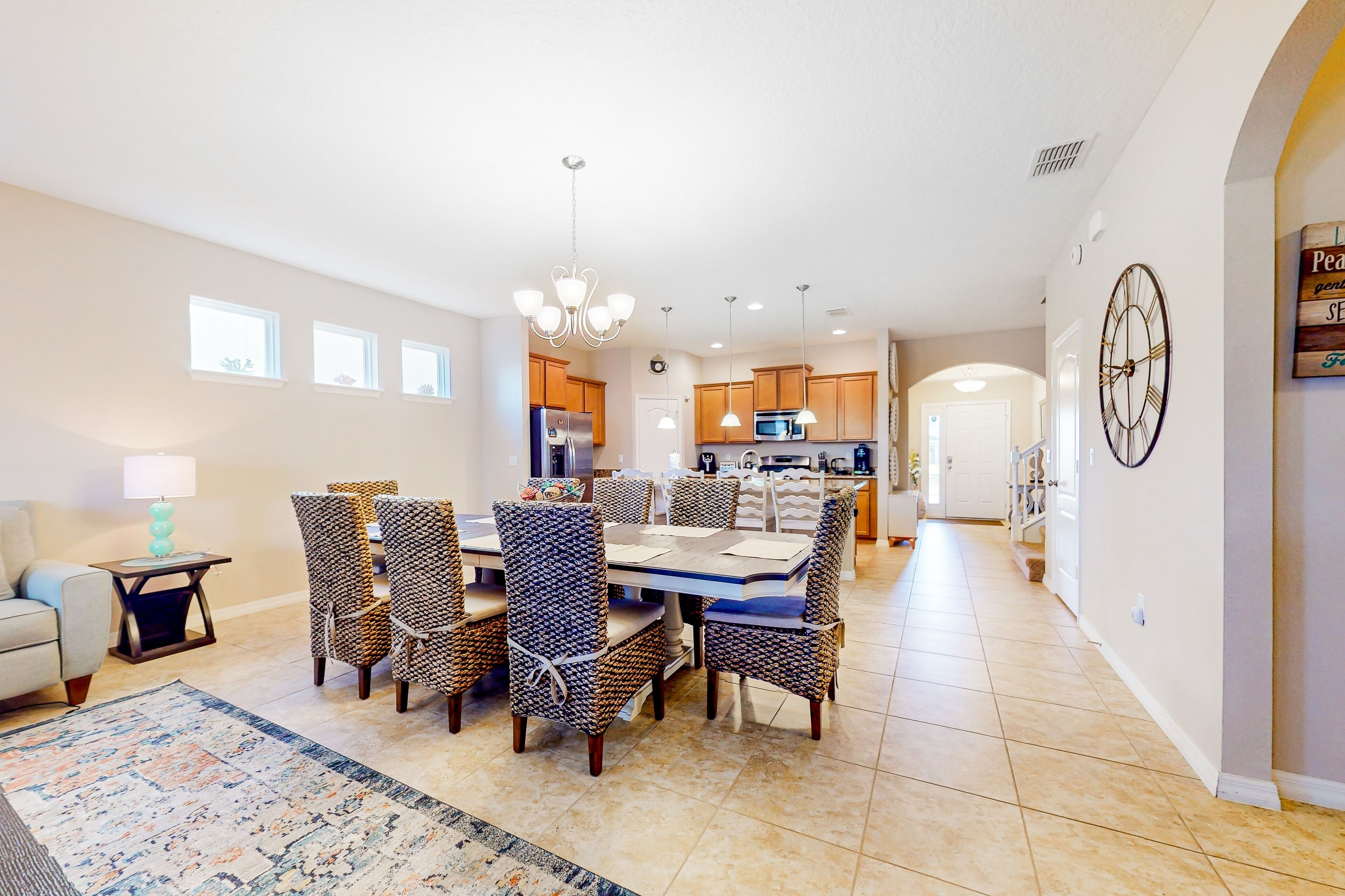 A spacious dining area with a large table and chairs, a kitchen in the background, and a clock on the wall.