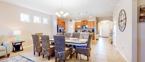 A spacious dining area with a large table and chairs, a kitchen in the background, and a clock on the wall.