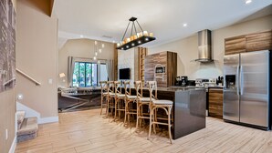 A modern kitchen with a central island, bar stools, stainless steel appliances, and a large window. - Modern villa with patio, grill, central AC, WiFi, shared pool, tennis, & gym (Kissimmee)
