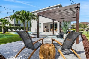 A patio area with two wicker chairs, a woven ottoman, and a pergola. - 8BR/7BA Kissimmee | Near Disney, Private Pool & Game Room (Kissimmee)