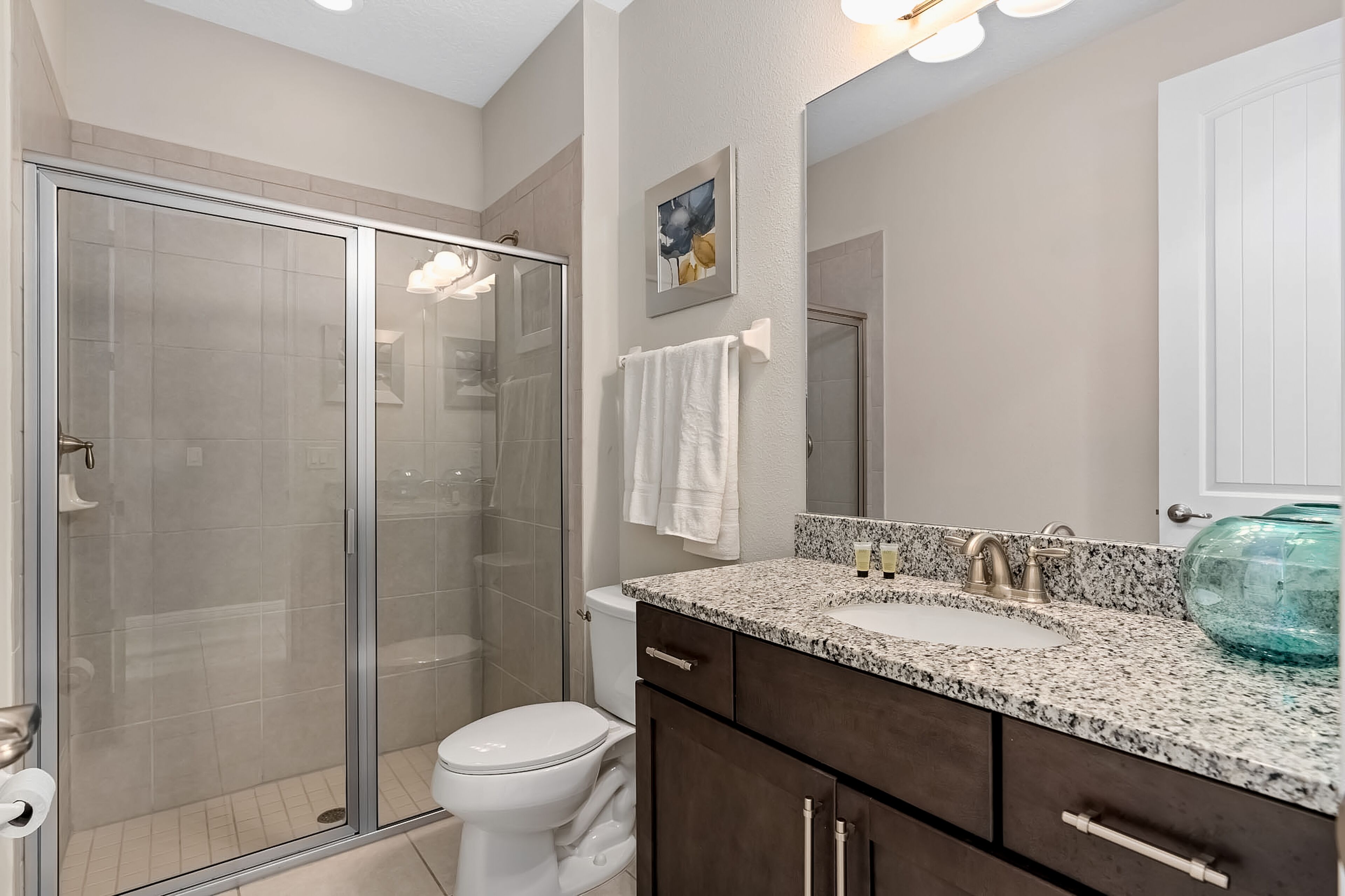 A modern bathroom with a glass shower enclosure, a white toilet, a granite countertop sink, and dark wooden cabinets.