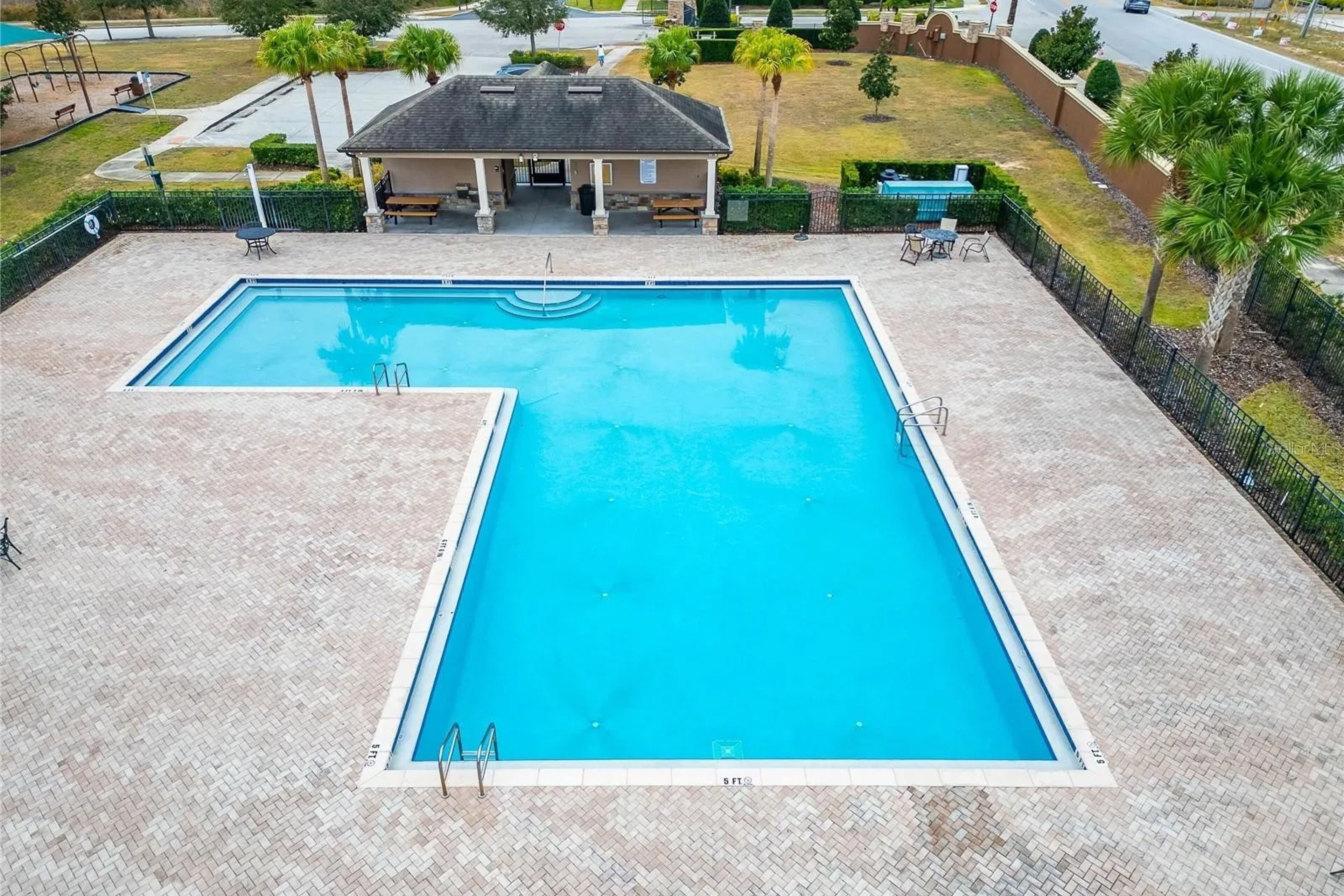 Une piscine rectangulaire avec une terrasse carrelée, entourée de palmiers et dotée d’un espace salon abrité.