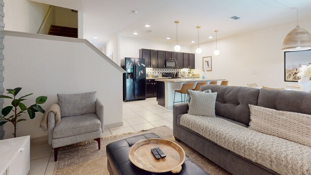 A modern living room with a grey sofa, a coffee table with a remote, and a staircase leading to a kitchen area. - Solterra Escape | Private Pool | Café, Tennis & Water Park with Lazy River (Davenport)