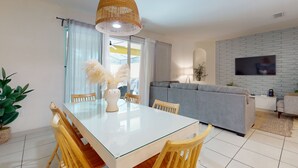 A modern living room with a glass dining table, wooden chairs, a grey sofa, and a TV mounted on a brick-patterned wall. - Solterra Escape | Private Pool | Café, Tennis & Water Park with Lazy River (Davenport)