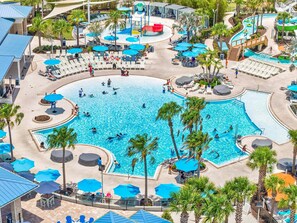 A large outdoor swimming pool surrounded by lounge chairs and umbrellas, with palm trees and a water slide in the background. - Exquisite home with pool, lanai, game room, central AC, & washer/dryer (Davenport)